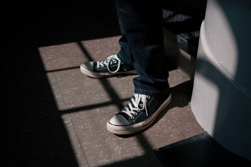 The bottom of a teenager's legs and his feet show as the boy stand at a window, wearing jeans and conversers.