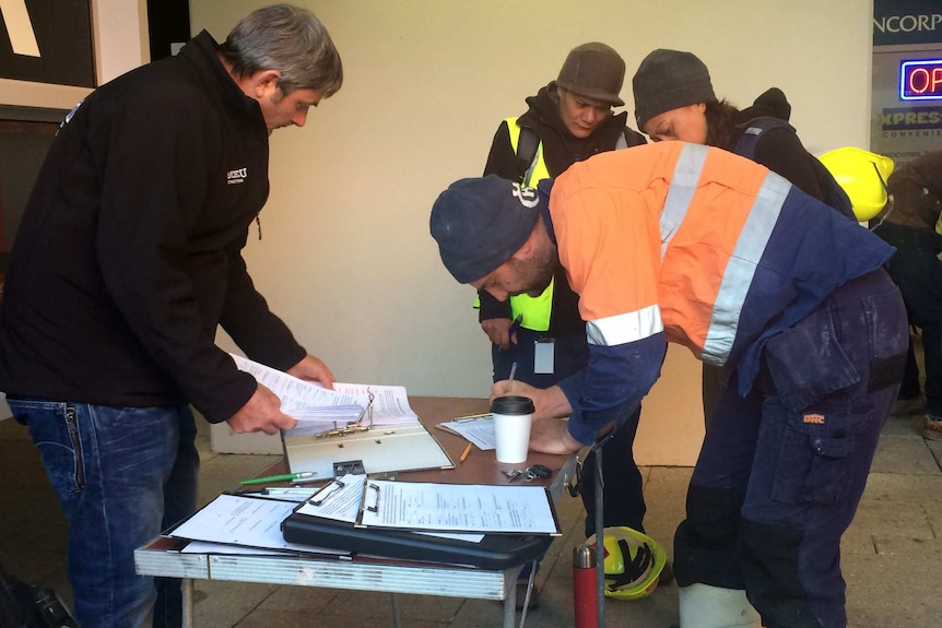 A worker in hi-vis clothing leans over a table signing a piece of paper as a man looks through a file folder opposite him.