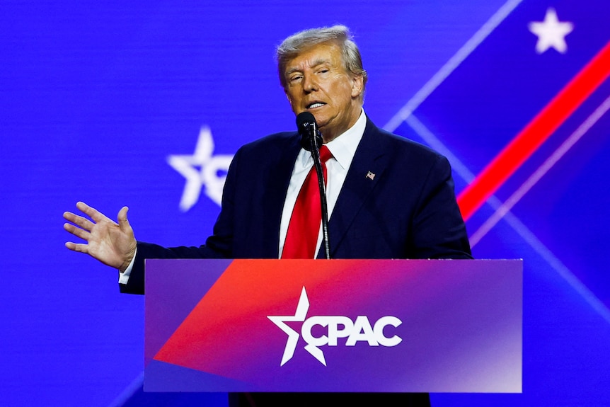 Donald Trump stands on stage behind a podium with CPAC written on it while wearing a dark suit with red tie.