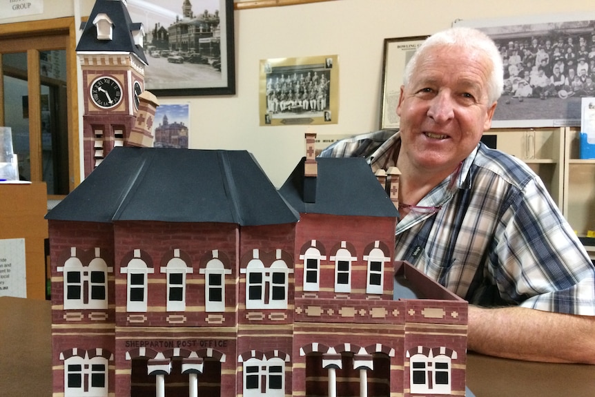 man with grey hair and check shirt sitting behind paper model of building