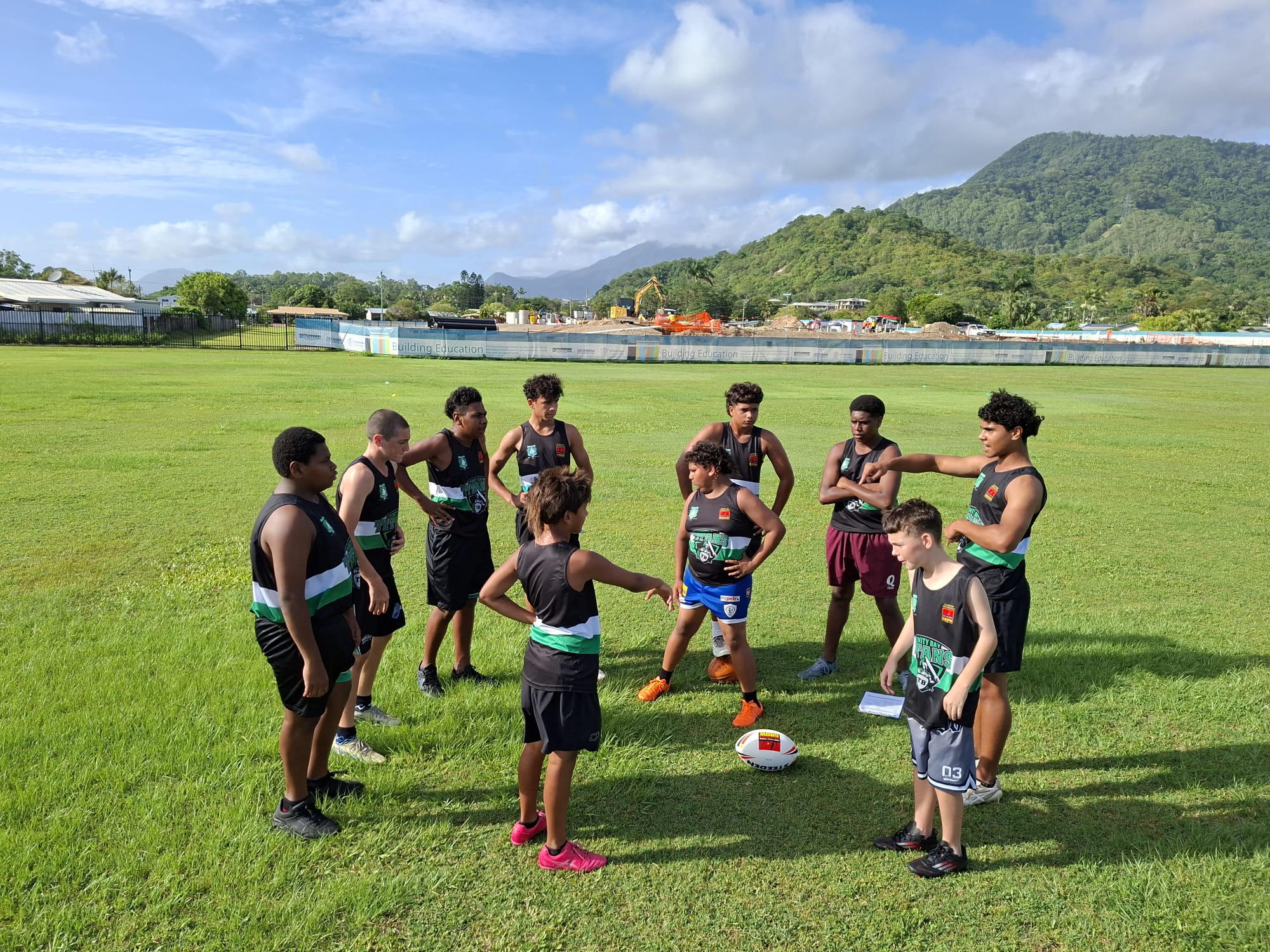 A group of Indigenous teenage boys wear sports uniform and stand in a circle on a grassy oval.