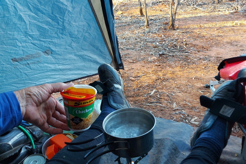 View from inside of Del preparing a noodle cup over a cooker, tent open, dirt and scrub outside