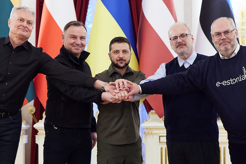 Five men pose in front of flags with their hands together in a circle