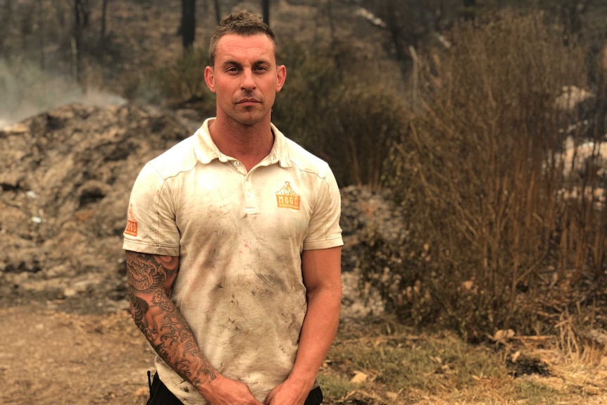 A man with a dirty shirt stands in front of bushfire affected land.