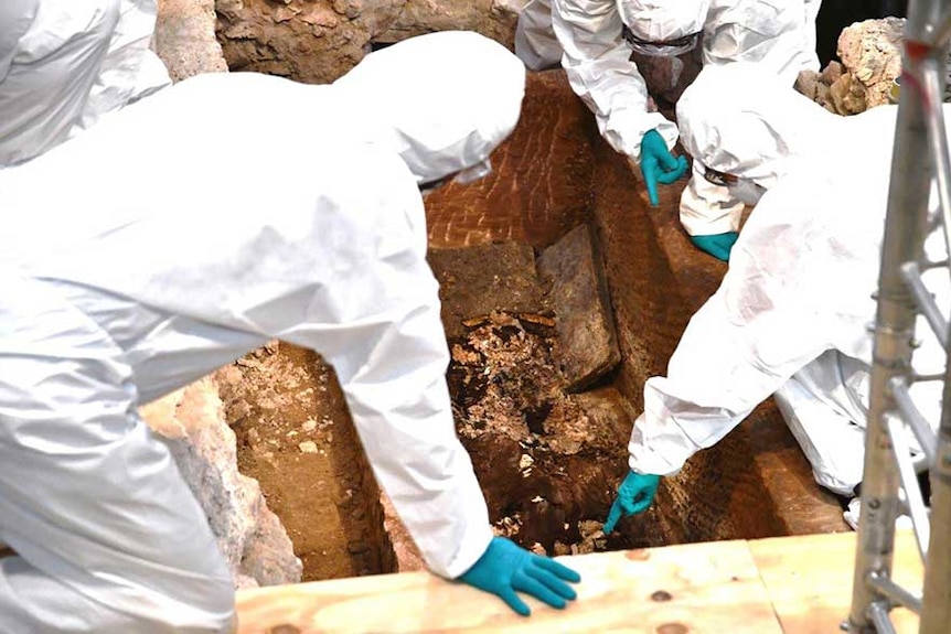 A group of people wearing gloves and protective clothing gathered around a sunken stone box.
