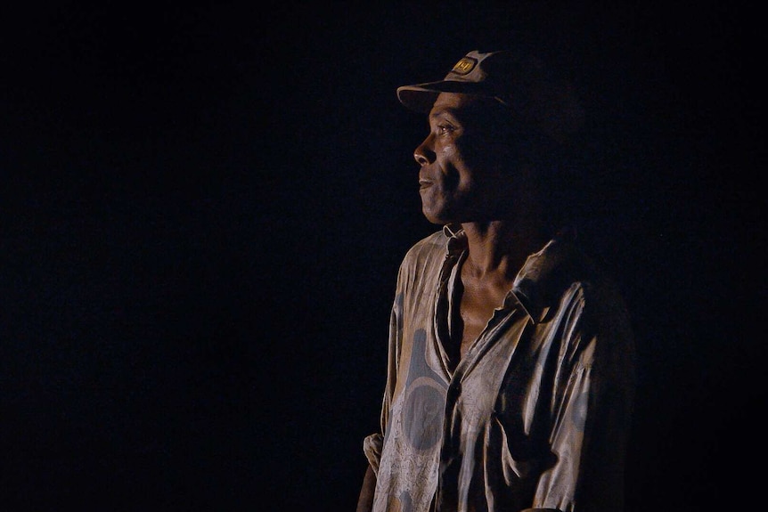 A man looks to the left of the camera with serious expression on face in image taken at night. He wears a cap and light shirt.