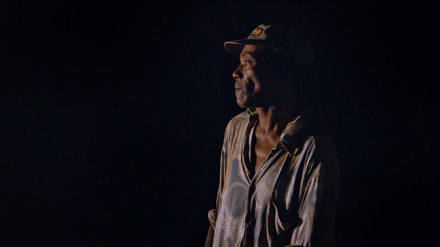 A man looks to the left of the camera with serious expression on face in image taken at night. He wears a cap and light shirt.