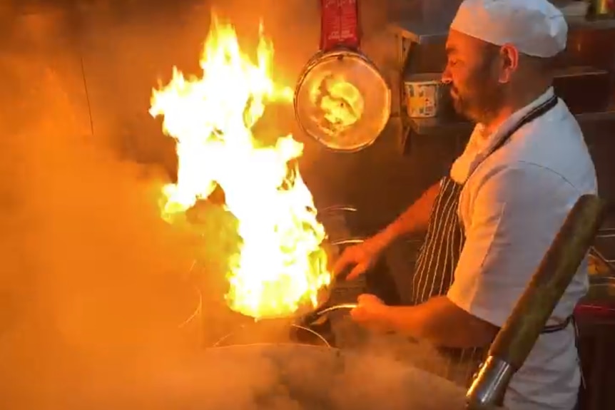 A chef sautee's vegetables in a pan. The oil makes bursts of flame rise into the air. A steaming vat of rice is also visible