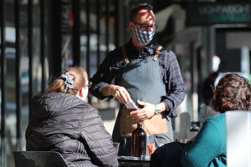 A male waiter in a facemask takes an order from two unidentifiable women seated at an outdoor cafe table.