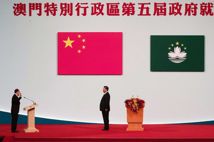 Chinese leader Xi Jinping stands in front of two large flags of China and Macau that hang on the wall.