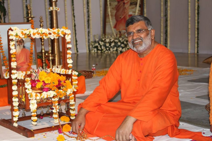 A man wearing saffron clothes sitting next to a flower-laden shrine smiles at the camera.