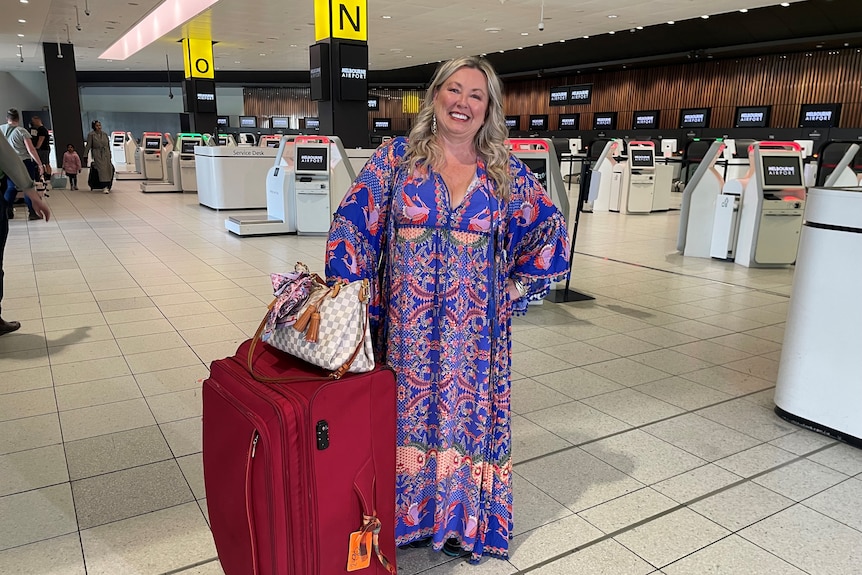 A blonde woman in a blue, red and yellow patterned dress standing at the airport with her suitcase.