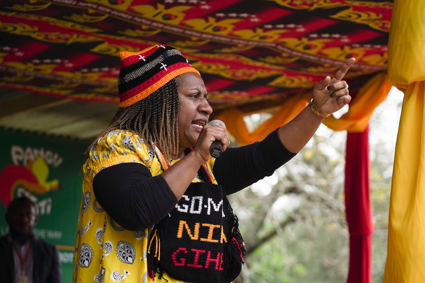 A woman in a yellow dress with black bag slung in front of her speaks into a microphone and points to the crowd