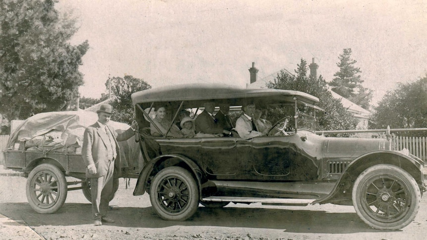 Black and white photo of a 1920s passenger truck with passengers and a trailer on the back full of cargo and mail sacks.