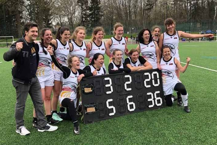 13 women pose for a photo on an oval dressed in football uniforms and holding a scorecard.