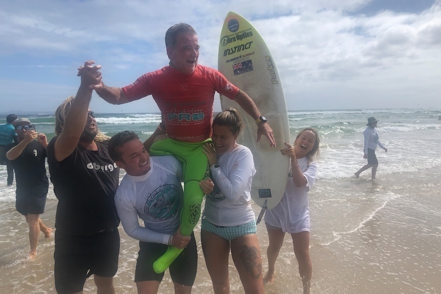 One-legged man in a yellow wetsuit and red rash vest is chaired on the shoulders of fans with his surfboard in the background.