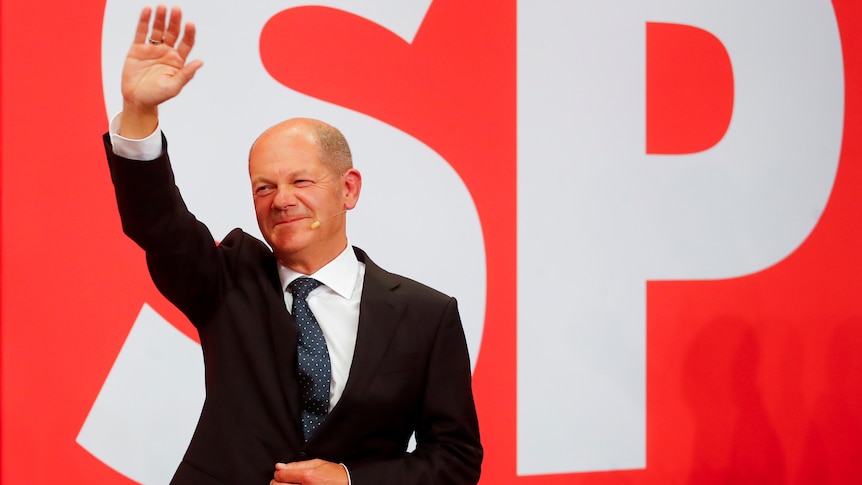 A man smiles and waves from in front of a red screen