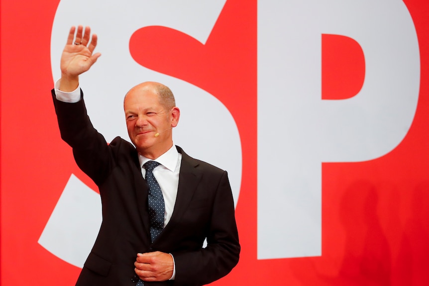 A man smiles and waves from in front of a red screen