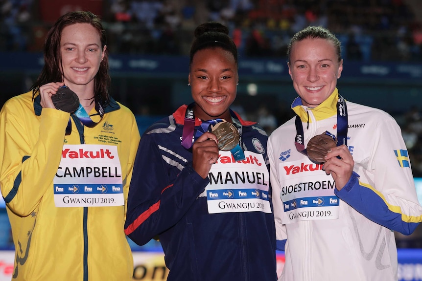 Three female swimmers stand holding their medals for photographers.