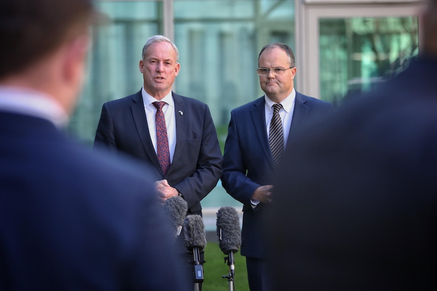 Two men in suits talk in front of a press pack at Parliament House