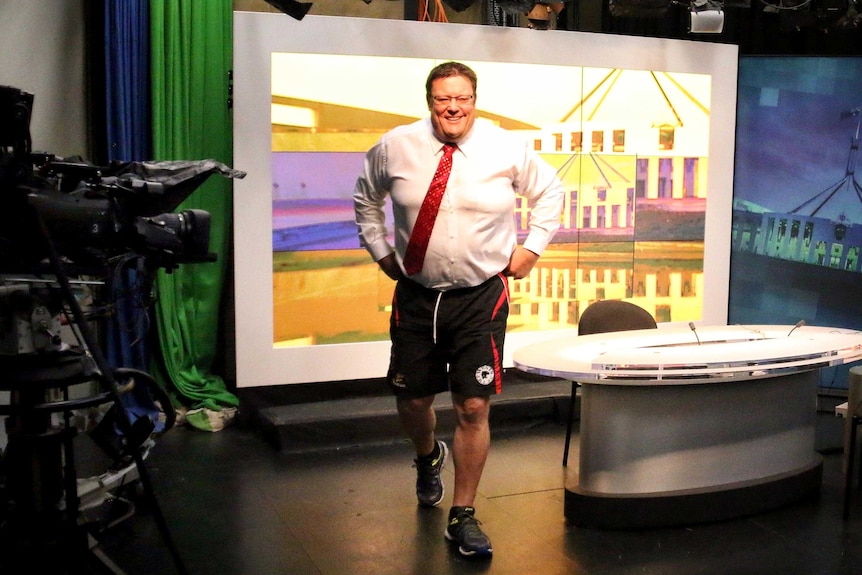 Glenn Lazarus walks through a TV studio in a shirt and tie but wearing shorts.