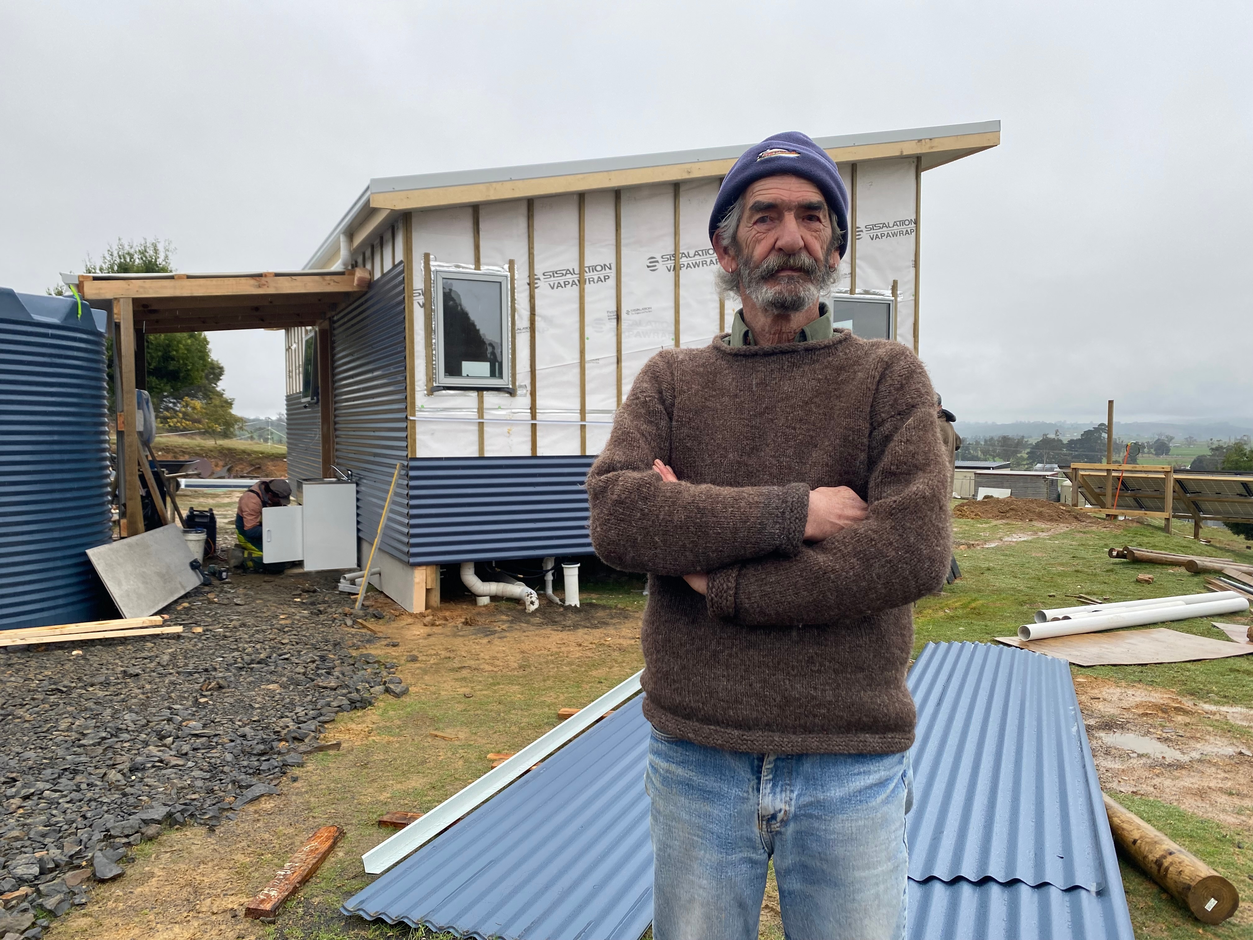 A man in his 70s wearing a beanie standing in front of a small home in construction.