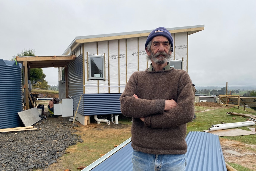 A man in his 70s wearing a beanie standing in front of a small home in construction.