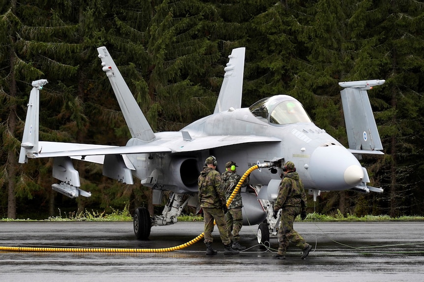three people in camouglage stand by a fighter jet with a yellowb pipe fueling it 