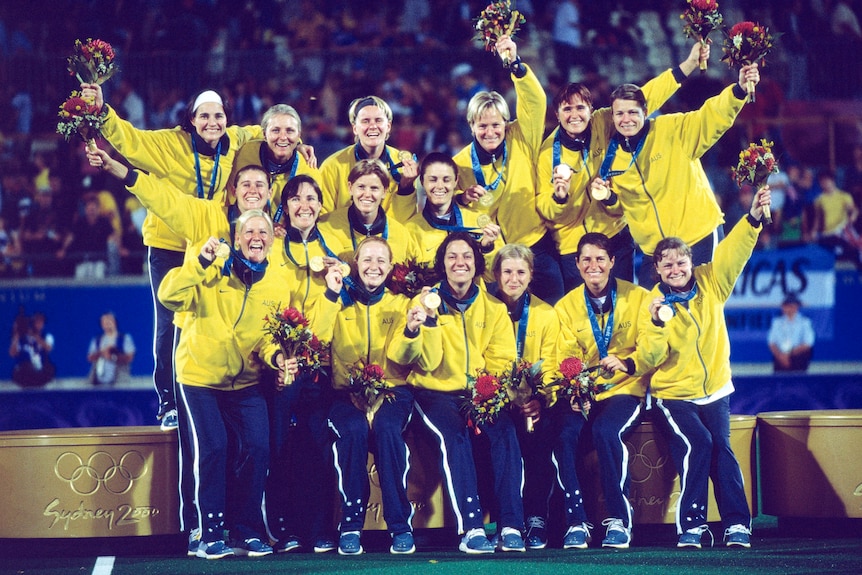 The Hockeyroos on the podium together, smiling, holding their gold medals and bouquets.