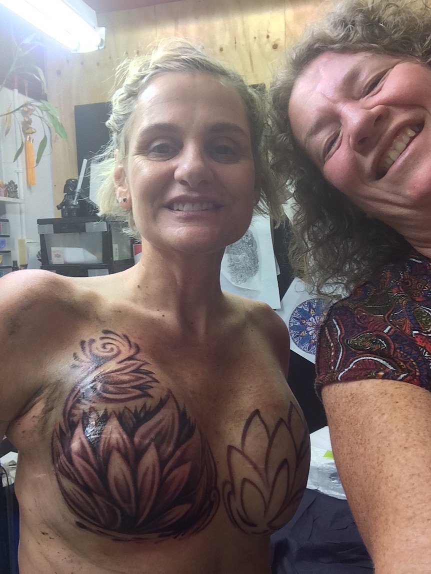 Two women smile for the camera, one of them naked with tattoos on her breasts.