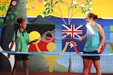 Indigenous girl in school uniform and Ash Barty tapping rackets in front of mural with Aboriginal and Australian flags