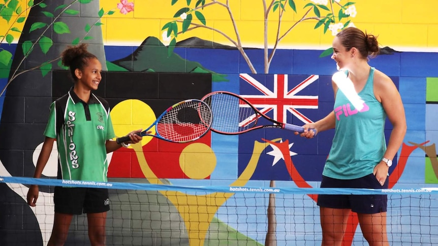 Indigenous girl in school uniform and Ash Barty tapping rackets in front of mural with Aboriginal and Australian flags