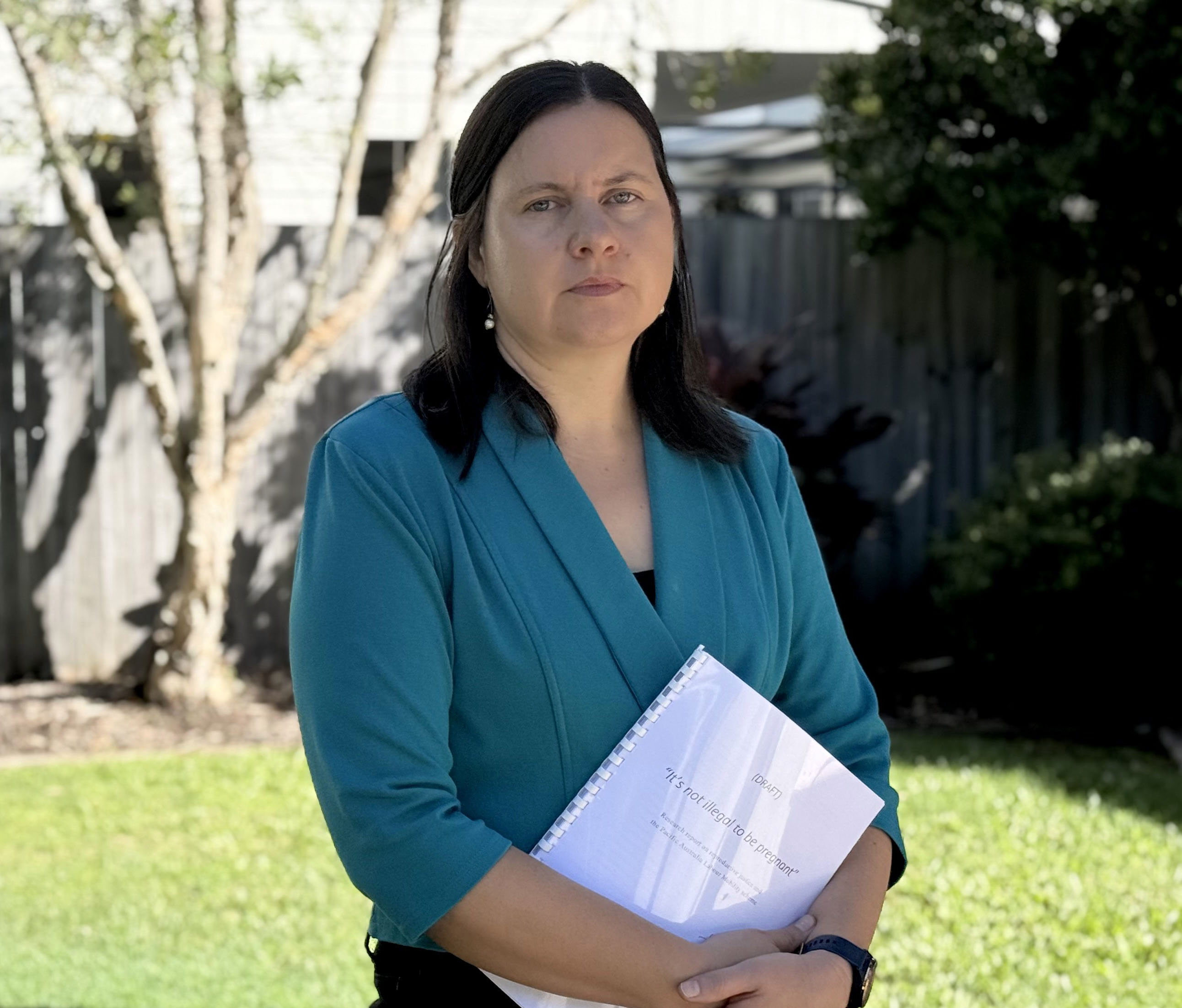A woman wearing a teal coloured suit jacket holds a report in her hands and looks ahead. She is standing a garden setting.