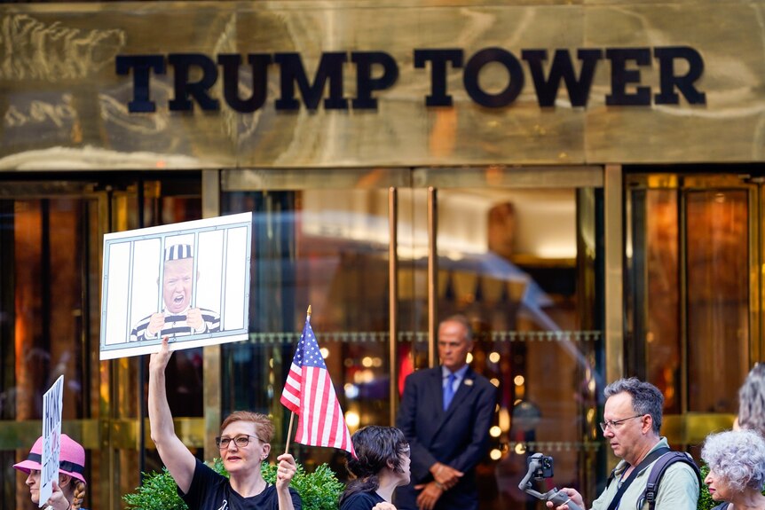 A crowd of people gather in front of the entrance to Trump Tower holding flags and a sign with Donald Trump behind bars.