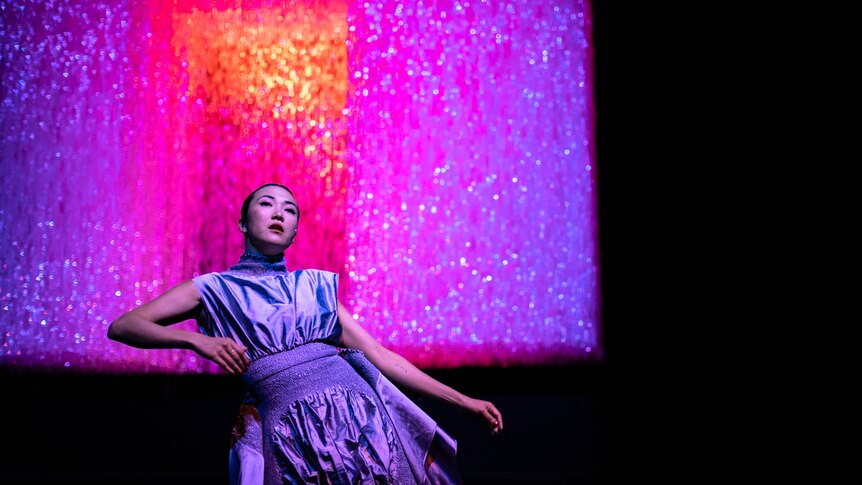 A Chinese Australian in her 30s with long dark hair wears a silver dress and performs in front of pink-lit beaded curtain.