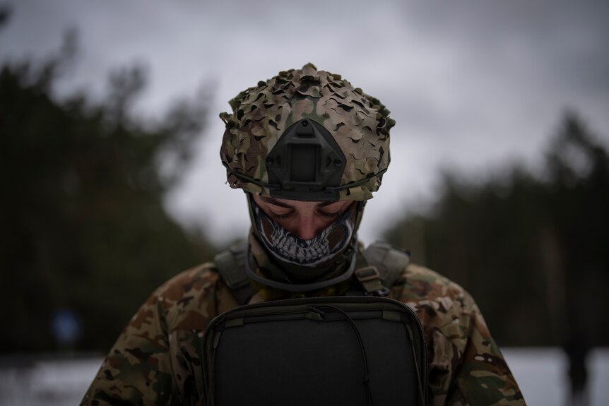 A soldier in military camouflage looks down. He has a mask covering half of his face. 