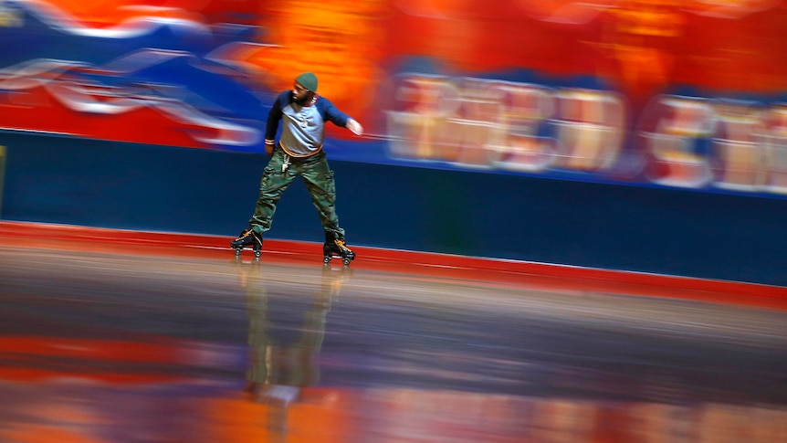 A man in green pants rollerskates with his feet apart along the inside of a rink. Bright colours blur behind him on the walls. 