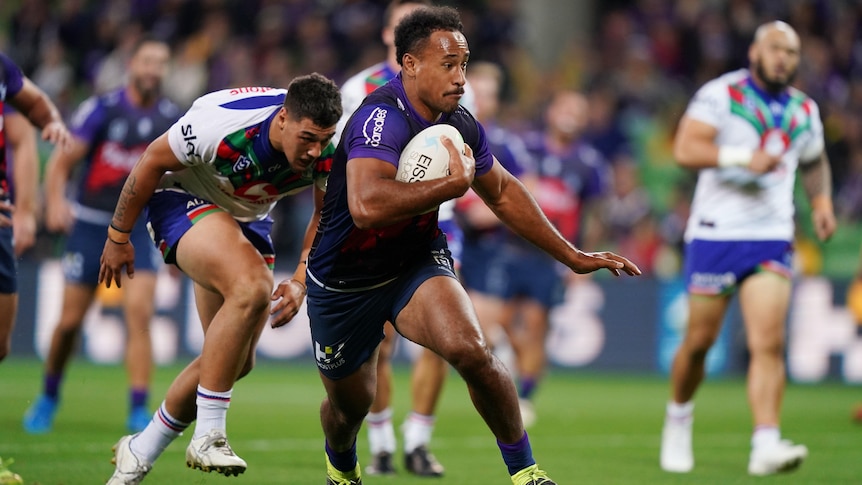 A Melbourne Storm NRL player runs with the ball held with his right arm against the Warriors.