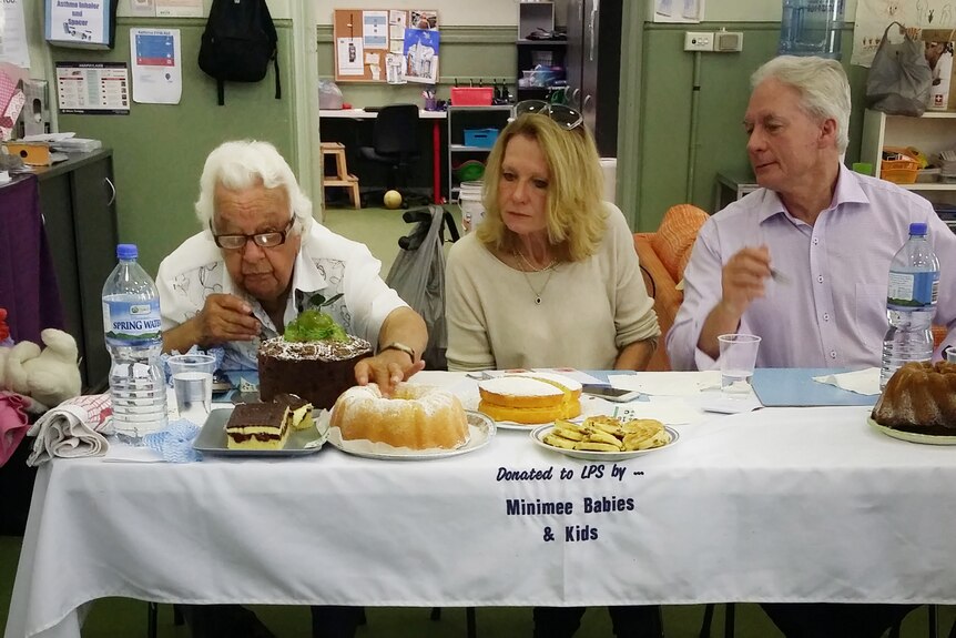 On the left, an older woman inspects a cake at a judging table with a woman and a man.
