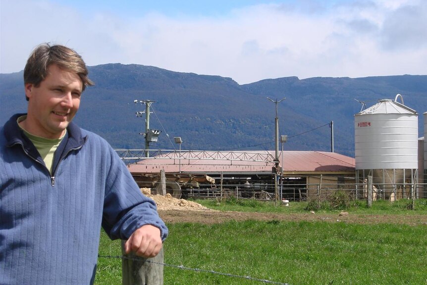 A man with short brown hair, wearing a blue jumper, leaning on a fence, with a milking shed in the background.