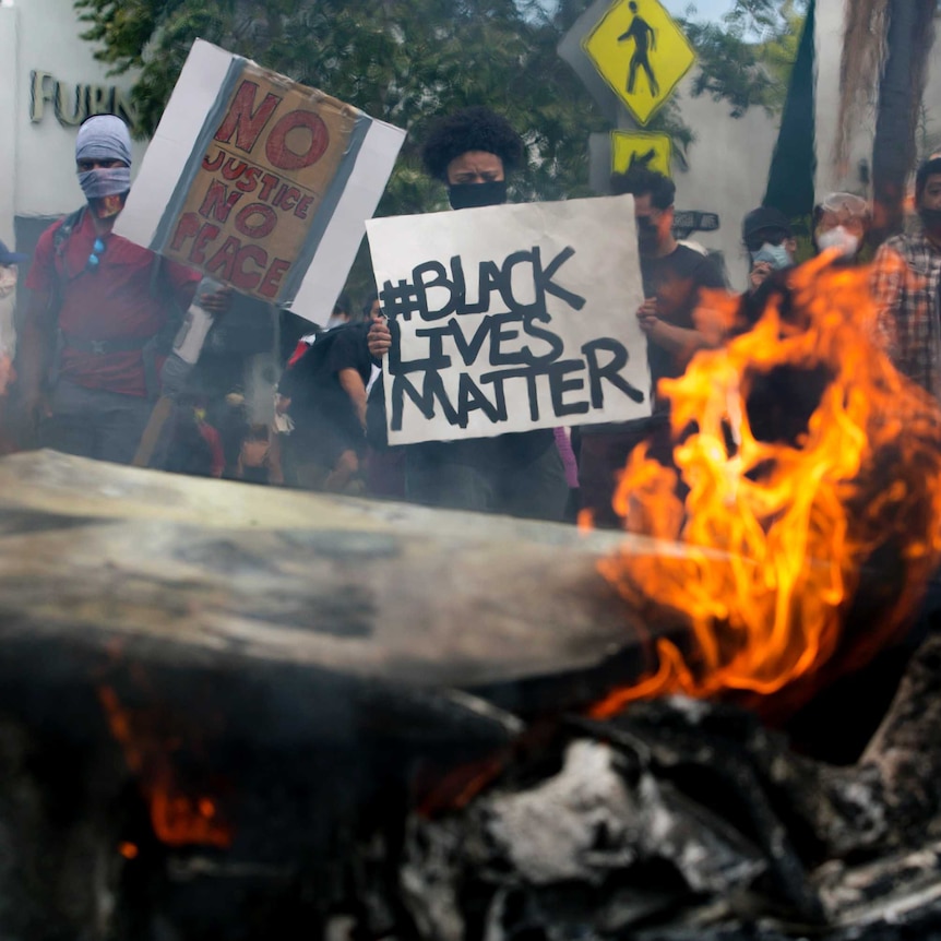 Protesters stand behind a burning car holding signs that read "Black Lives Matter" and "No Justice No Peace".