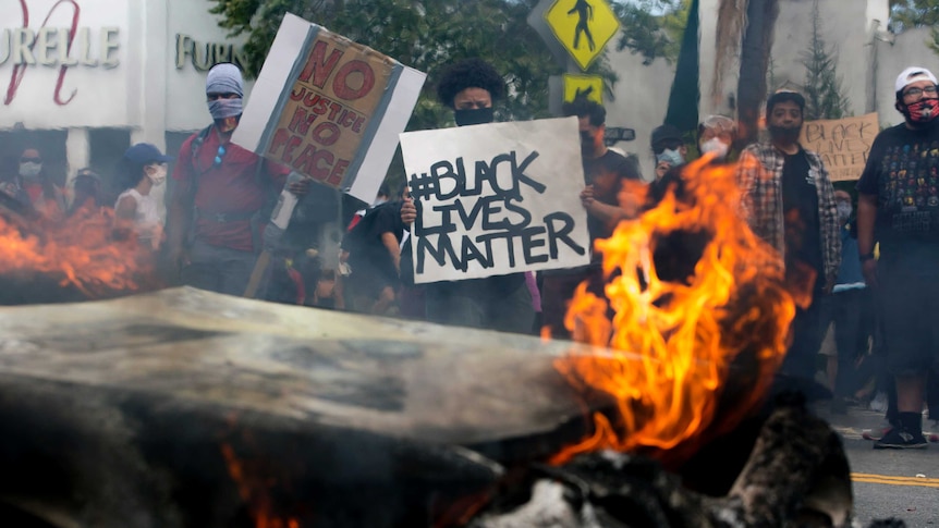 Protesters stand behind a burning car holding signs that read "Black Lives Matter" and "No Justice No Peace".