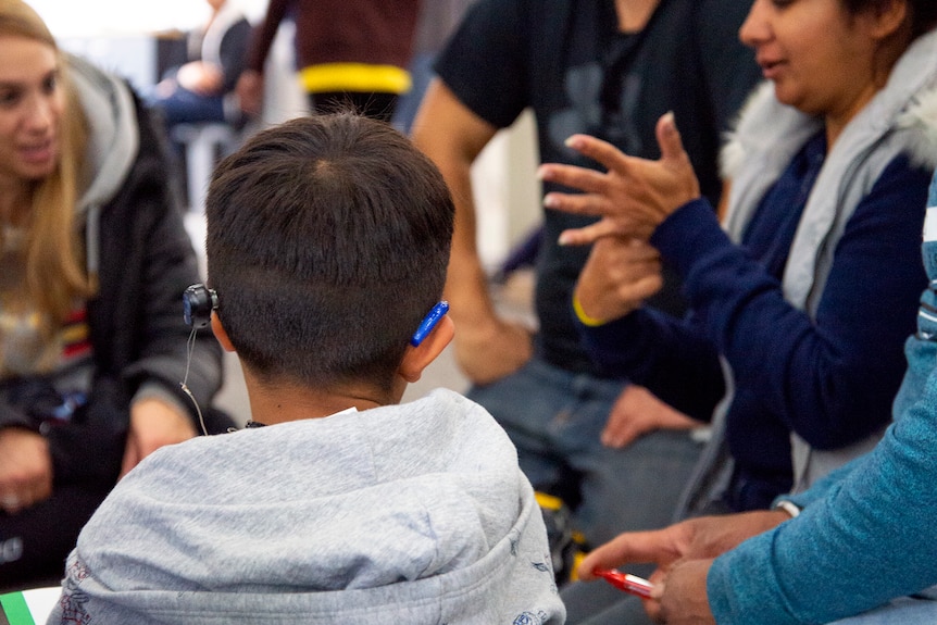 An image of the back of a child with cochlear implant and hearing aid looking at a woman signing 