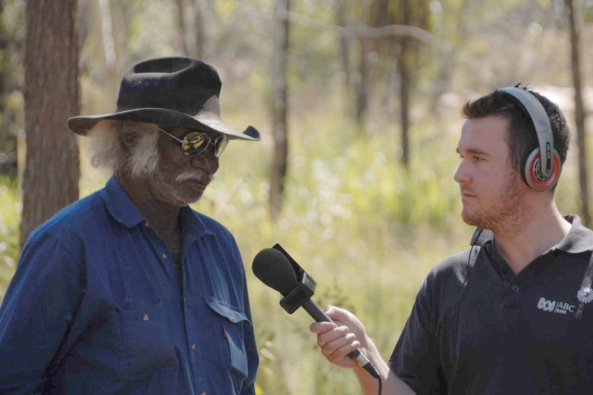 A journalist holding a microphone interviews a man in a bush hat and aviator sunglasses in the bush.