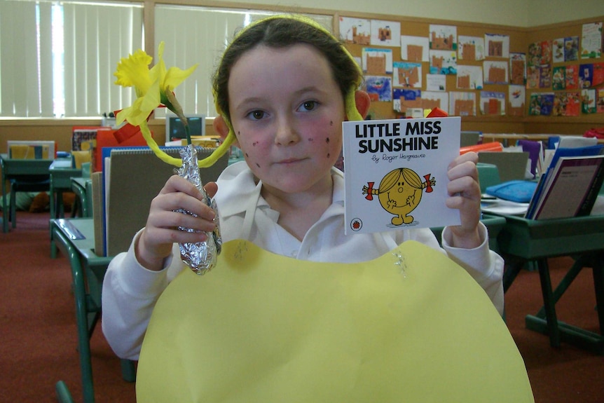 Findlay wears a white shirt and has yellow paper draped over herself. She holds a book and a daffodil and has a yellow headband.
