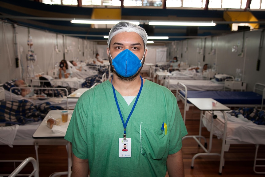 A young male nurse in green scrubs, a blue mask and white cap standing in a hospital ward