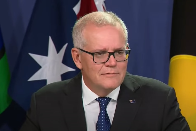 A man with short grey hair wearing black thin-framed glasses and a suit speaks in front of two flags.