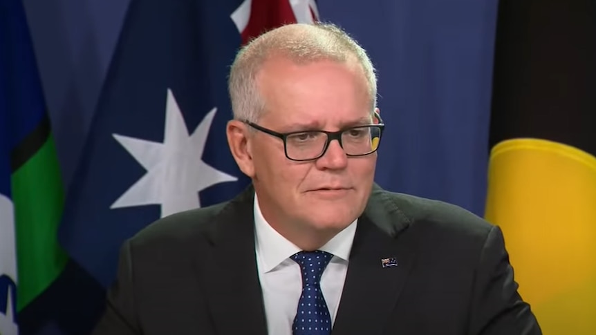 A man with short grey hair wearing black thin framed glasses and a suit speaking in front of two flags