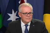 A man with short grey hair wearing black thin framed glasses and a suit speaking in front of two flags