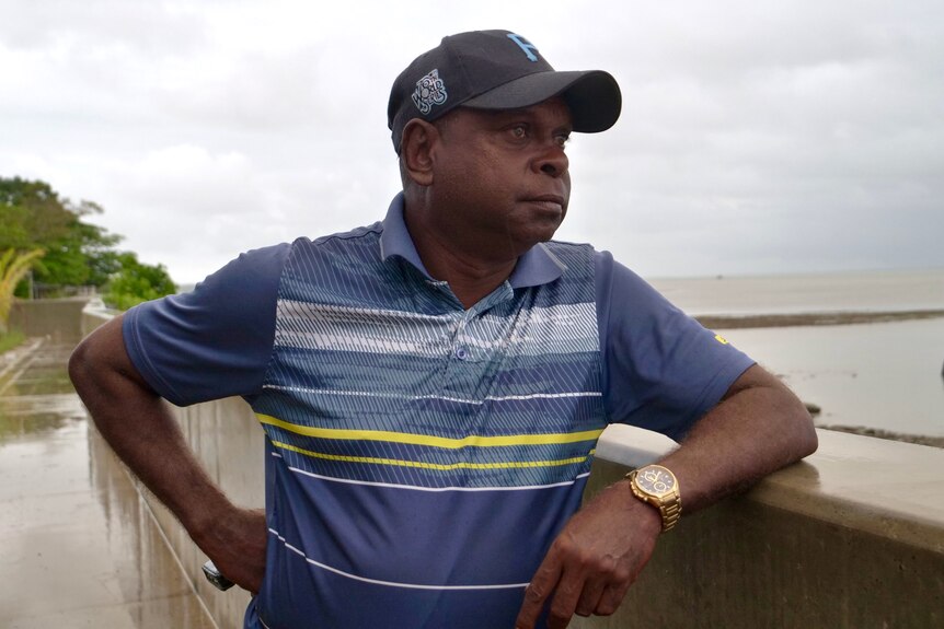 A man wearing a blue and yellow polo and a dark cap leaning against a wall looking out to sea